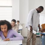 Estudantes fazem uma prova em sala de aula enquanto uma professora os supervisiona.