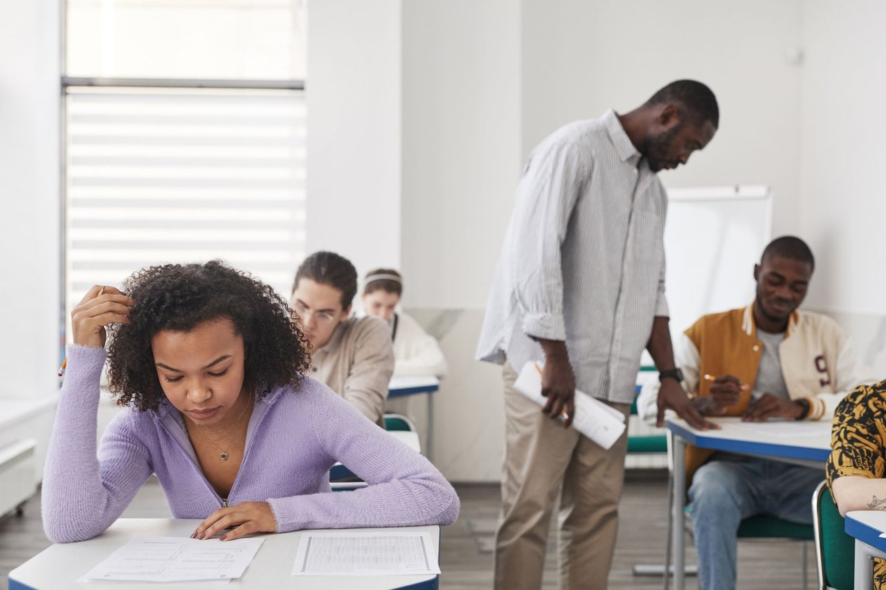 Estudantes fazem uma prova em sala de aula enquanto uma professora os supervisiona.