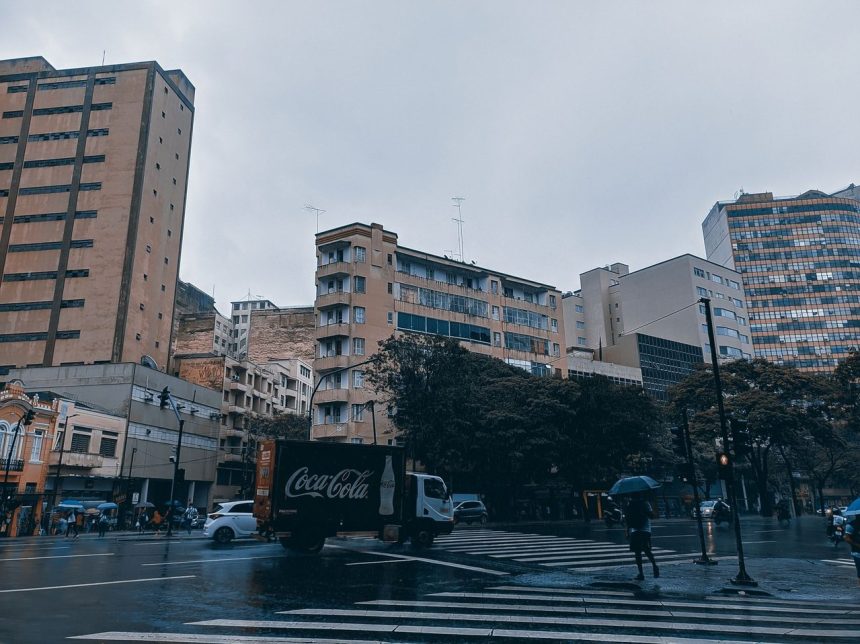 Pedestres e veículos atravessam uma rua movimentada de Belo Horizonte durante uma chuva intensa.