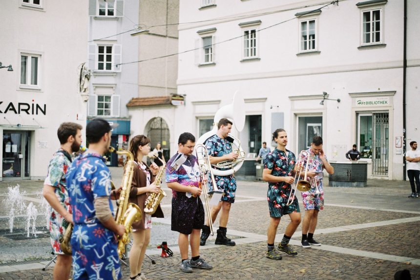 Banda de rua se apresenta com instrumentos de sopro em uma praça urbana, diante do público.
