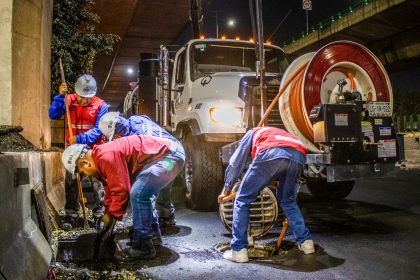 Trabalhadores fazem a limpeza de uma rede de drenagem urbana com máquinas, à noite, sob um viaduto.