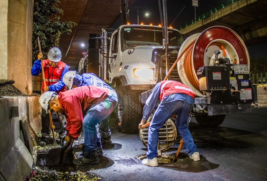 Trabalhadores fazem a limpeza de uma rede de drenagem urbana com máquinas, à noite, sob um viaduto.