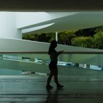 Silhueta de uma mulher caminhando em um espaço arquitetônico moderno enquanto consulta o celular.
