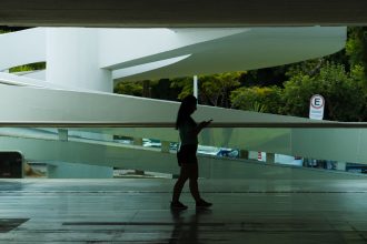 Silhueta de uma mulher caminhando em um espaço arquitetônico moderno enquanto consulta o celular.