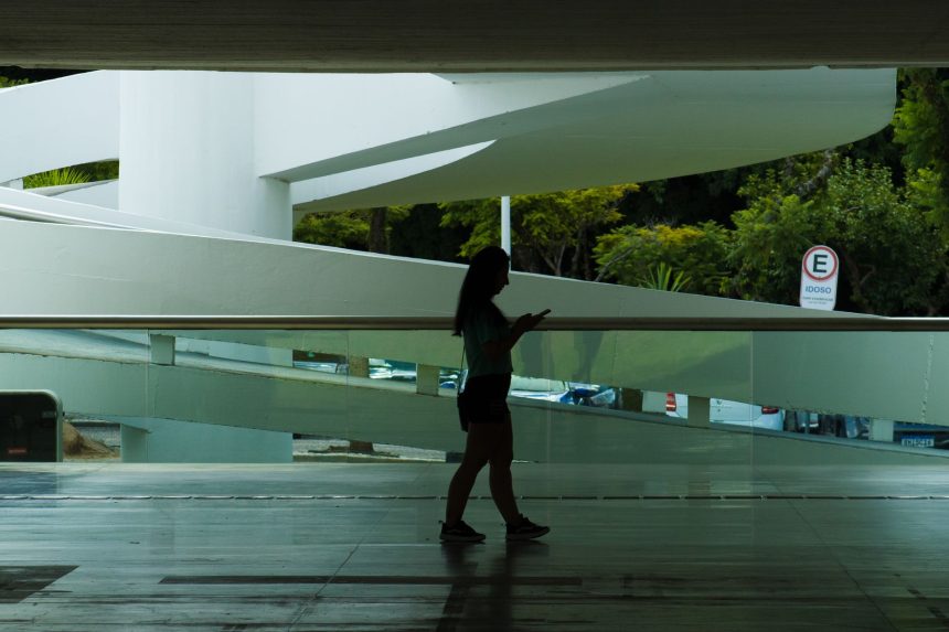 Silhueta de uma mulher caminhando em um espaço arquitetônico moderno enquanto consulta o celular.
